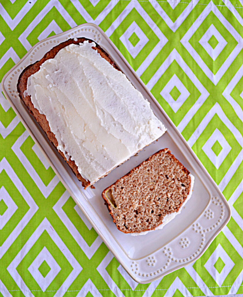 A white platter with banana bread topped with frosting and a slice cut from it. 