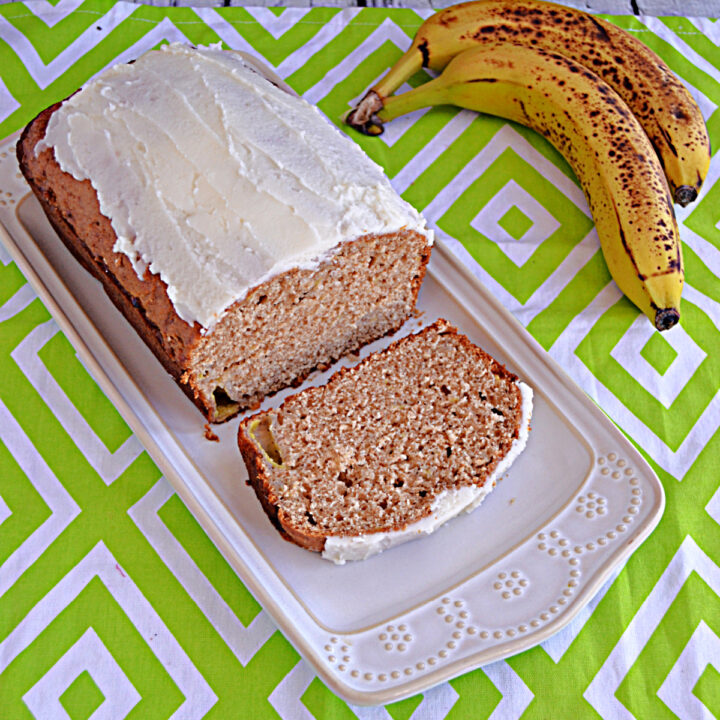 A white platter with a loaf of banana bread with a slice cut off and a bunch of bananas behind the platter.