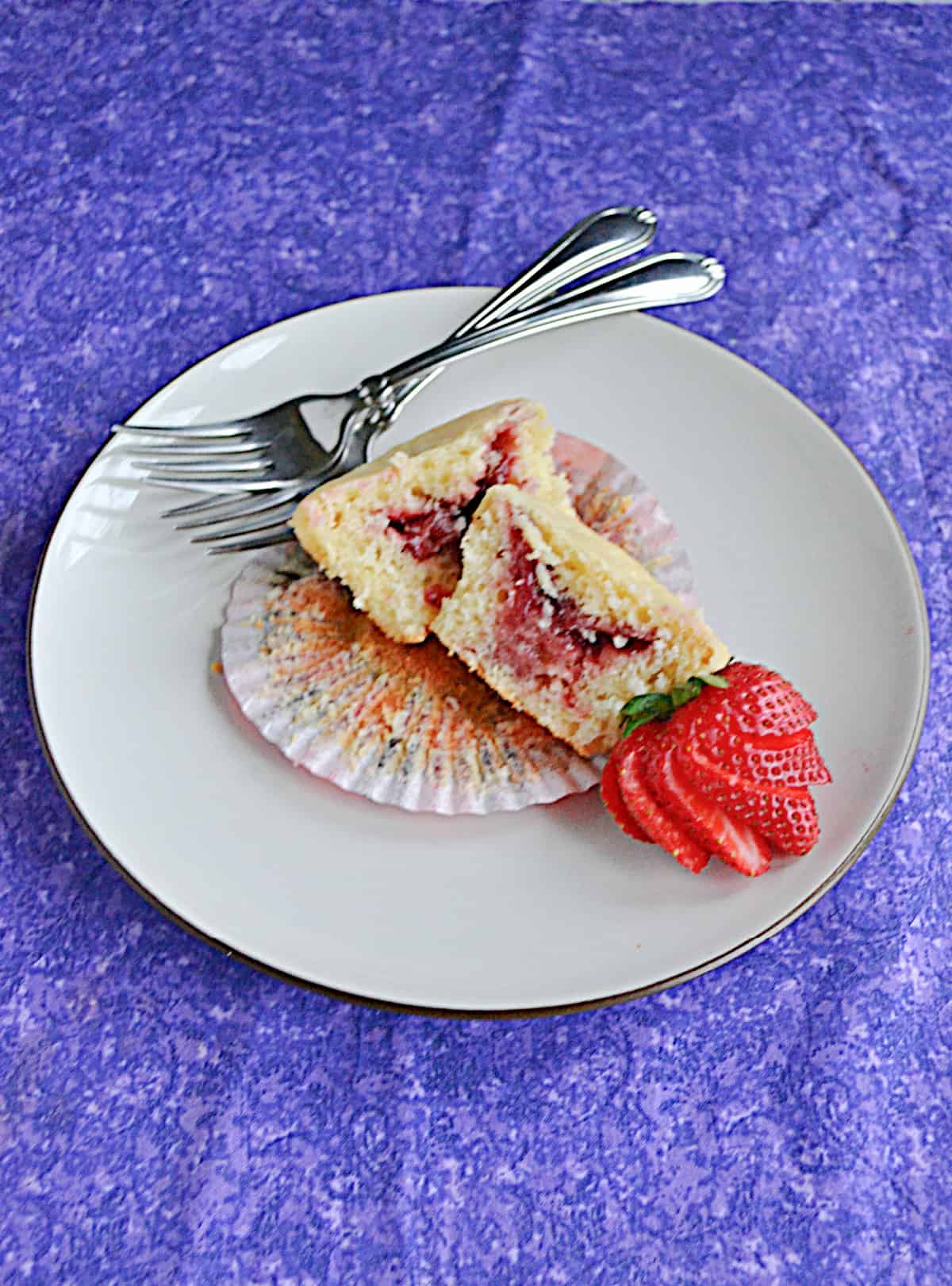 A plate with a muffin sliced in half with strawberry jam showing in the middle, a sliced strawberry, and two forks. 