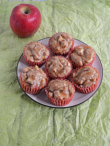 A plate with 7 apple cupcakes topped with brown sugar on it and an apple behind the plate.