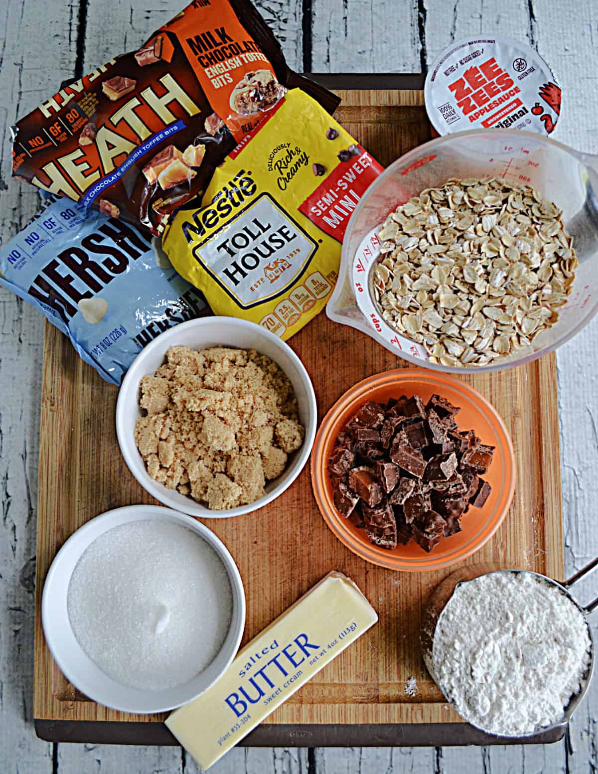 A wooden cutting board with a bag of Heath chips, white chocolate chips, and chocolate chips, a cup of oats, applesauce, sugar, and a stick of butter.