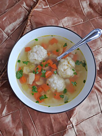 A bowl of Chicken and DUmpling Soup with three big dumplings, carrots, and celery, with a spoon in it.