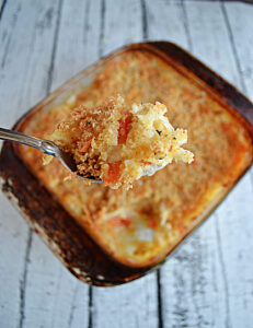 A close up of a spoonful of hash brown casserole with a baking dish of the casserole in the background.