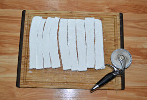 A sheet of homemade marshmallows that are cut into strips with a pizza cutter beside them.