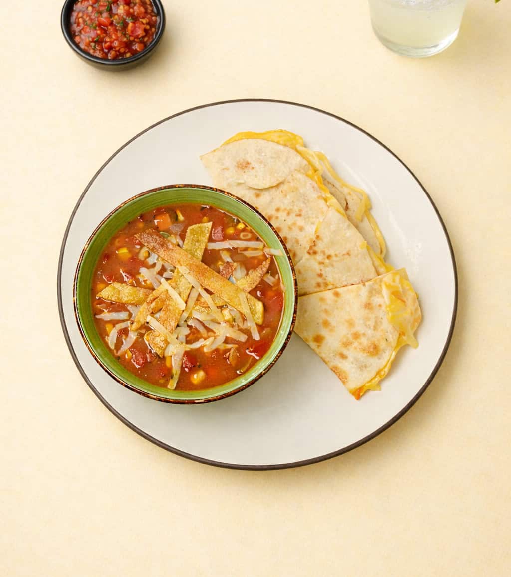 A plate with a bowl of soup on it and a quesadilla.