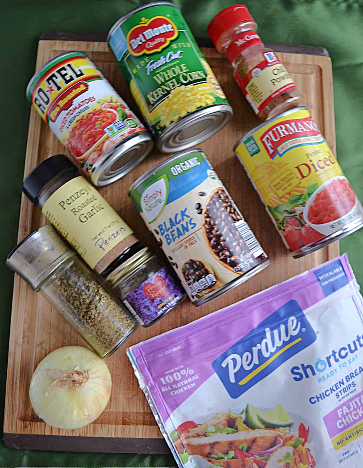 Wooden cutting board with spices, cans of food, chicken strips, and an onion on it.