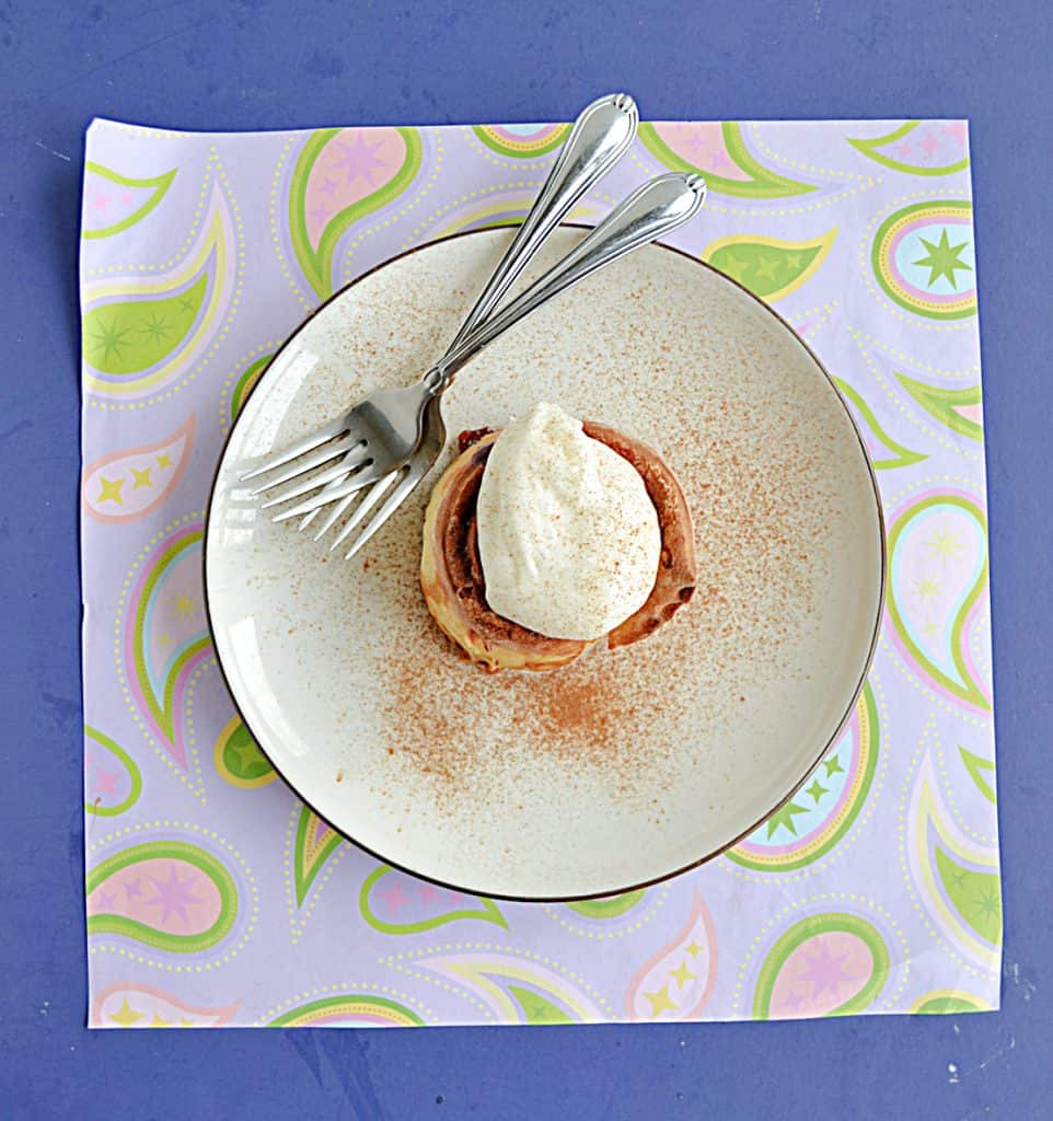 A plate with a cinnamon roll topped with frosting and two forks on the plate. 