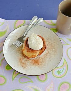 A plate with a cinnamon roll topped with frosting and two forks on the plate and a coffee cup behind the plate.