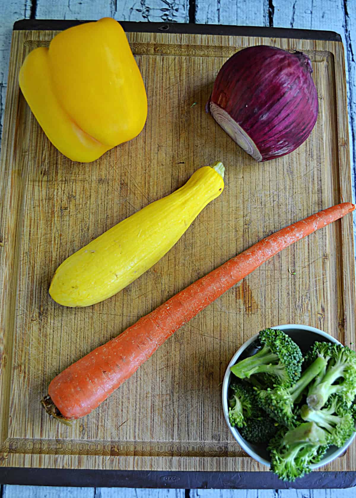 A cutting board with a pepper, an onion, a carrot, broccoli, and a squash on it.