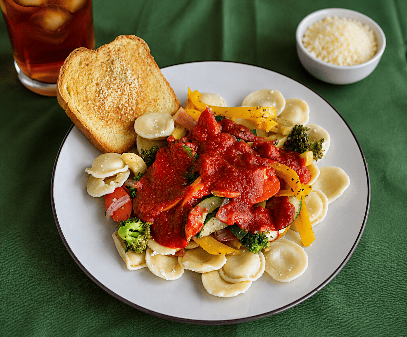 A plate of ravioli and vegetables with sauce and a piece of garlic bread, a bowl of parmesan cheese and a glass of iced tea behind the plate.