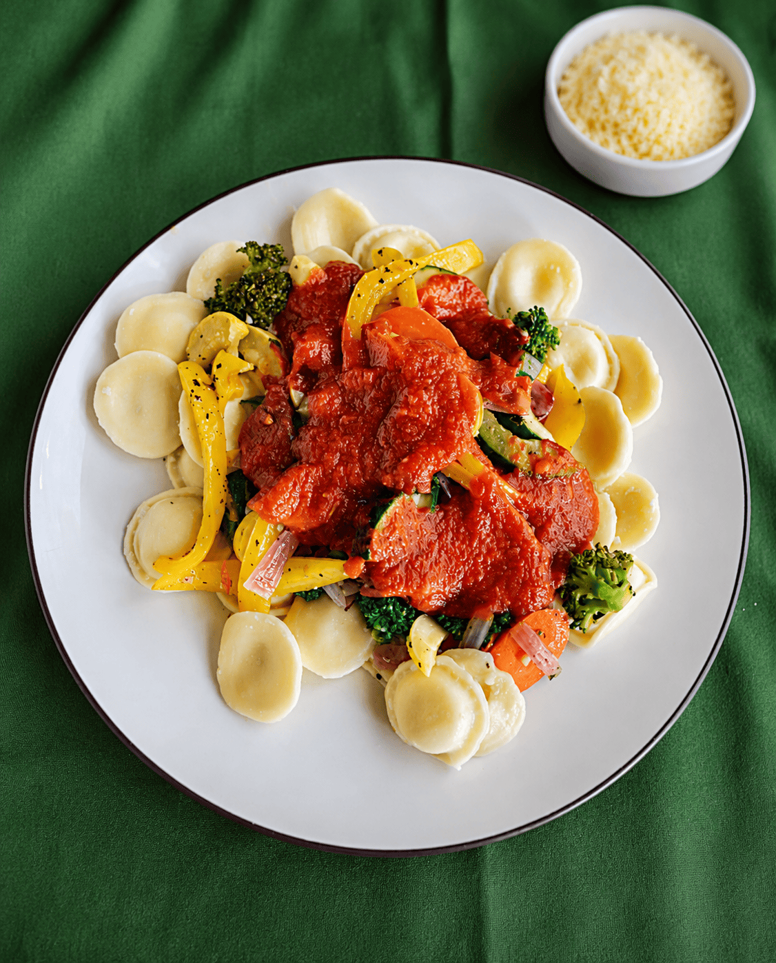 A plate of ravioli and vegetables with a small bowl of Parmesan cheese behind the plate.