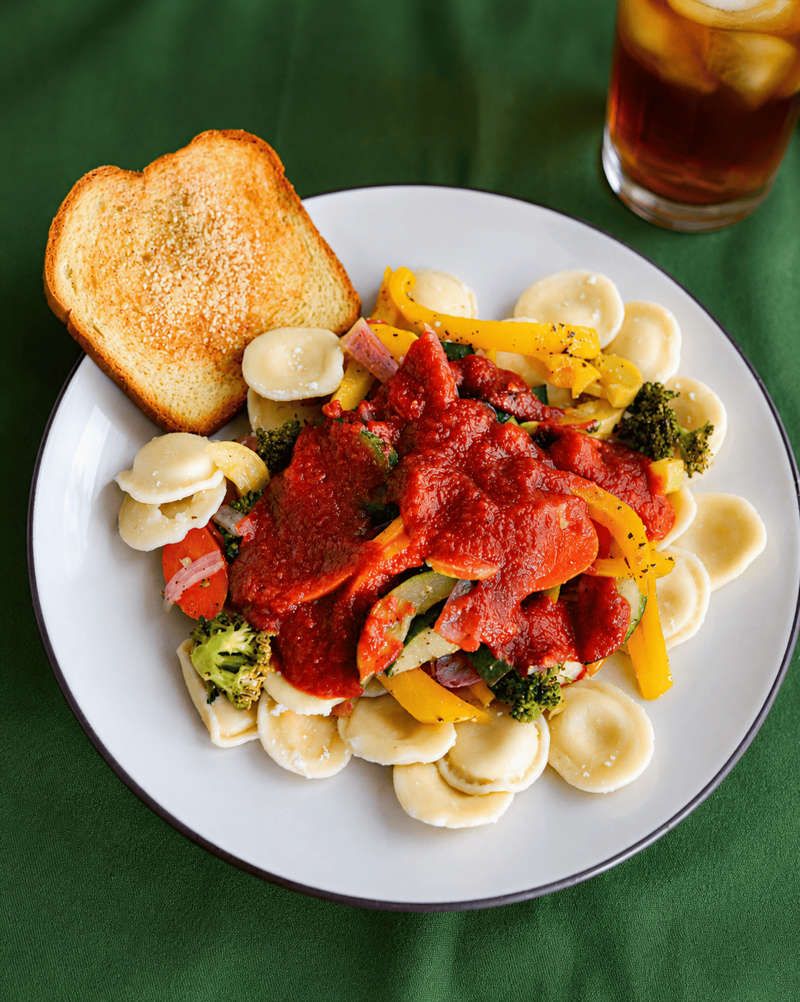 A plate of ravioli and vegetables with sauce and a piece of garlic bread and a glass of iced tea behind the plate.