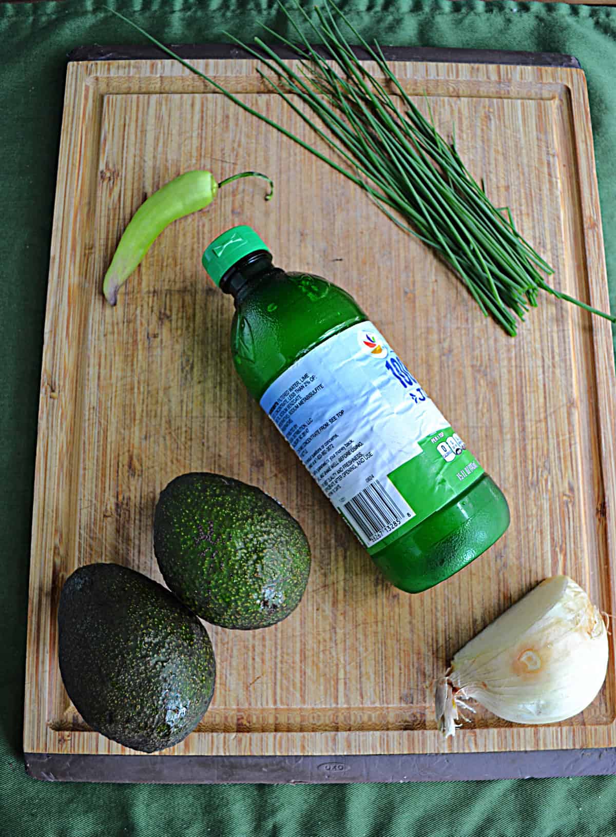 A cutting board with avocados, lime juice, an onion, chives, and a pepper on it.