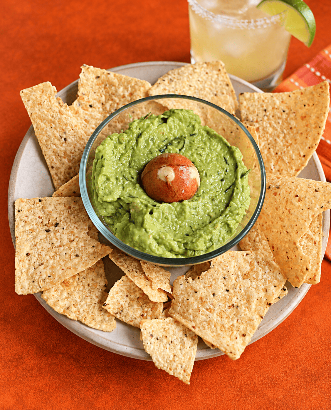 A plate of tortilla chips with a bowl of guacamole in the middle and a margarita behind the plate.