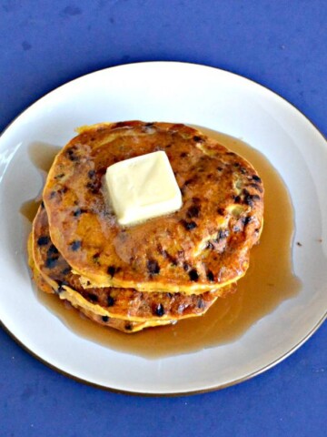 A white plate topped with a stack of three orange colored pumpkin pancakes studded with chocolate chips with a large pat of butter in the center and covered in syrup on a blue background.