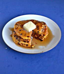 A white plate with a stack of three orange pumpkin pancakes studded with chocolate chips with a triangular cut out of it on a blue background.