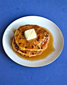 A white plate topped with a stack of three orange colored pumpkin pancakes studded with chocolate chips with a large pat of butter in the center and covered in syrup on a blue background.