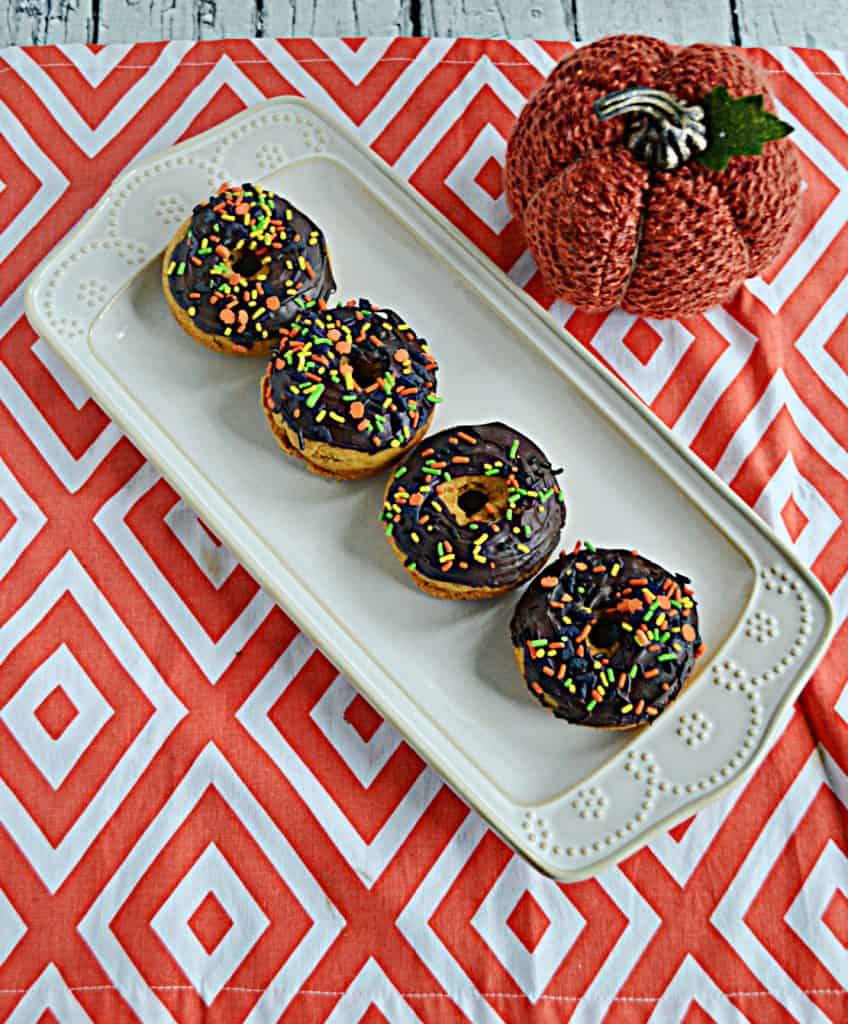 A platteer with four pumpkin donuts topped with chocolate glaze and a pumpkin behind them.