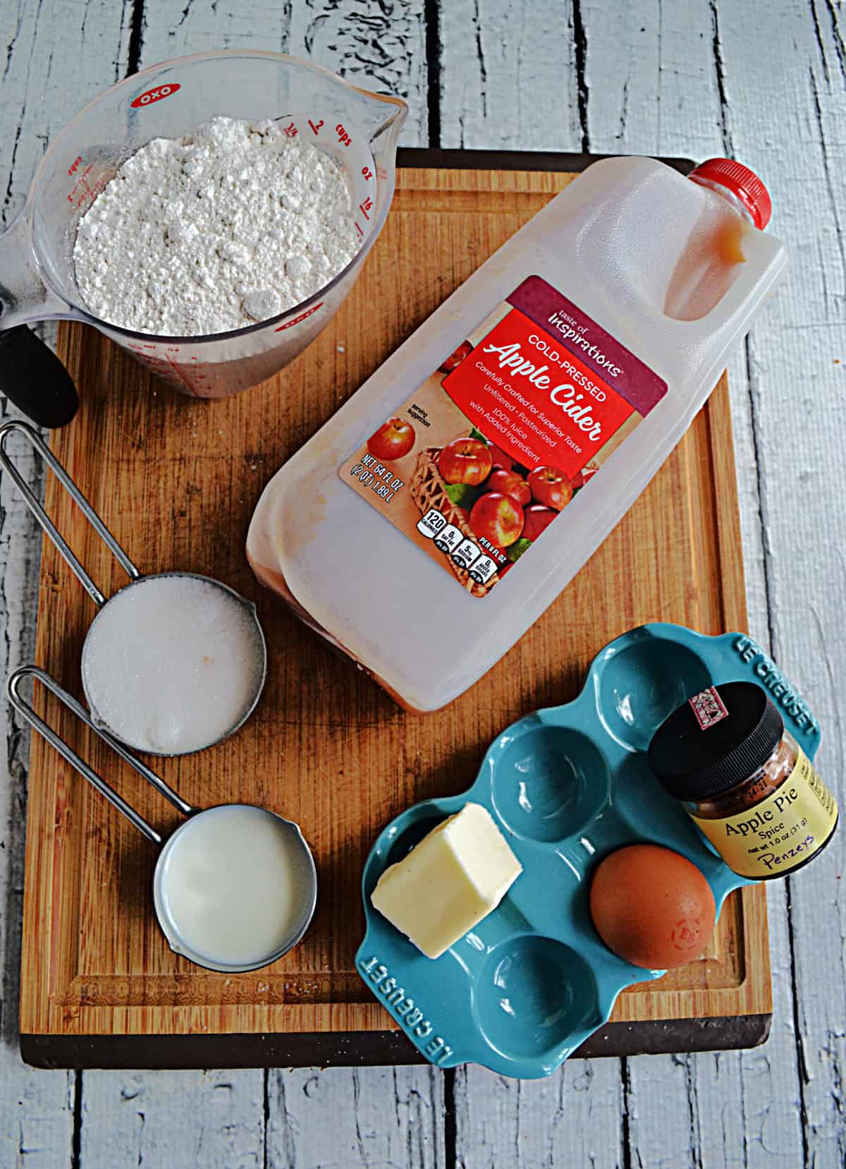 A cutting board with a carton of apple cider, an egg, a cup of flour, a cup of sugar, and spice jars on it.