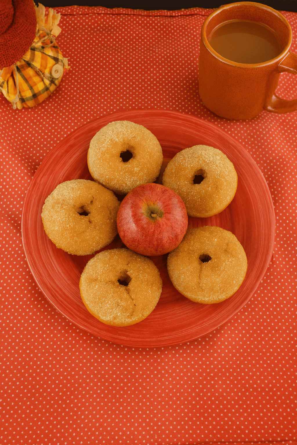 A plate of donuts with an apple in the middle and a cup of cider behind the plate.