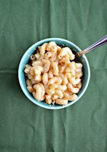 A small bowl filled with macaroni and cheese with a fork stuck in it from the top left on a green background.