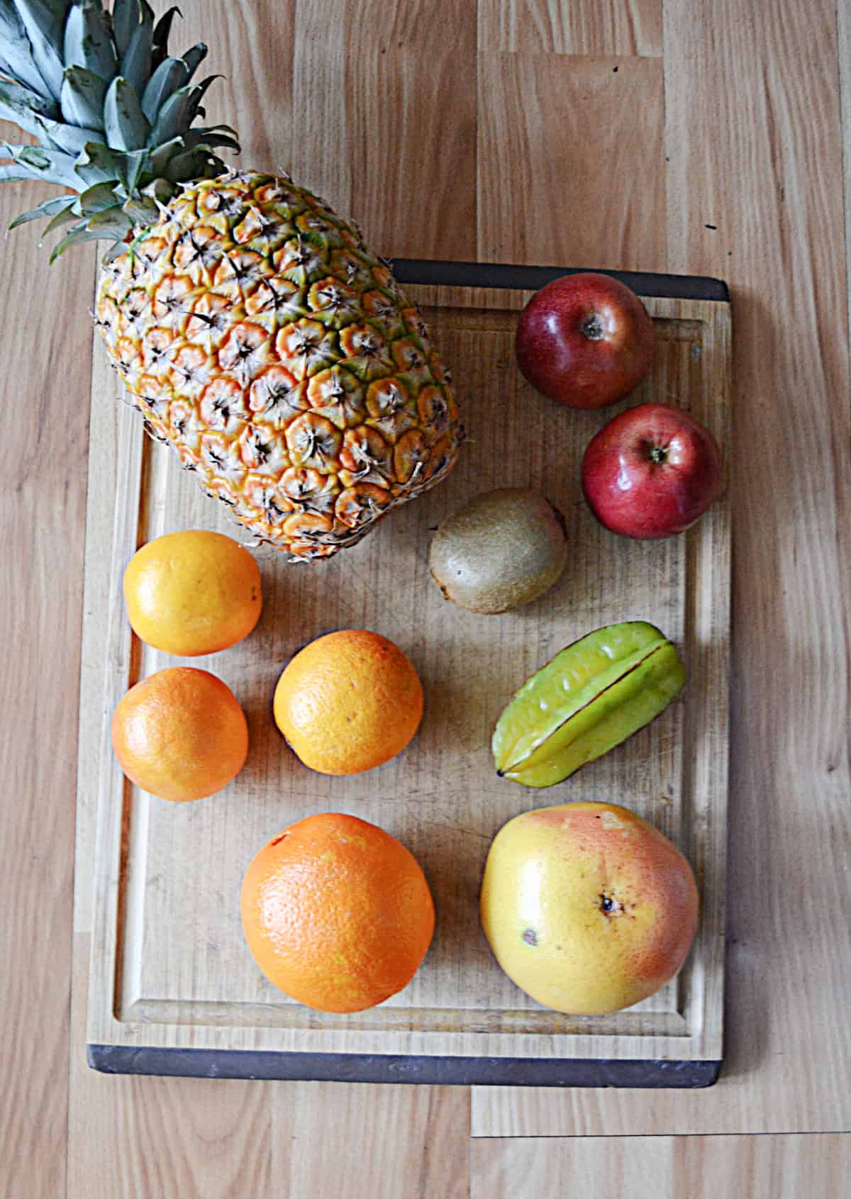A wooden cutting board with a pineapple, apples, oranges, grapefruit, kiwi, and star fruit.