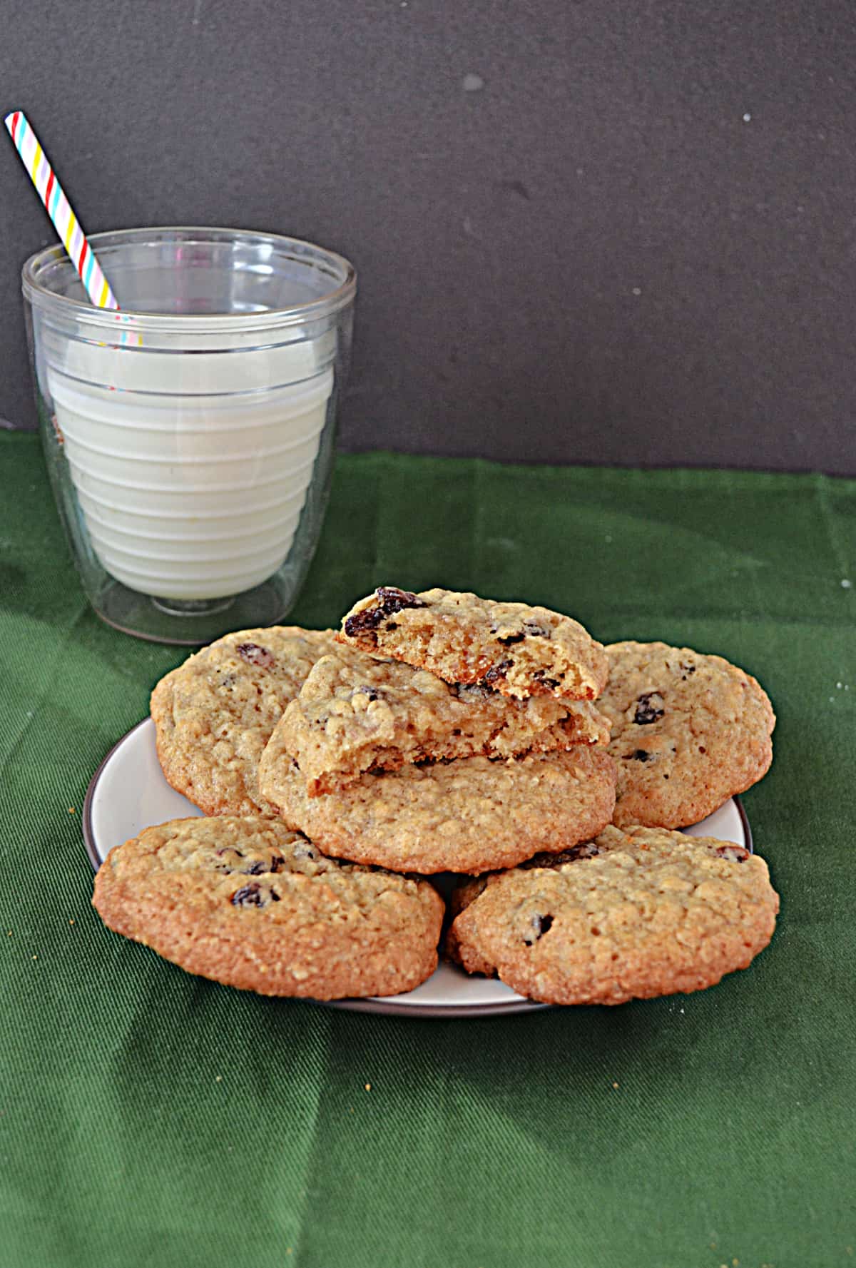 A plate of oatmeal raisin cookies with a cookie broke in half and a glass of milk behind the plate.