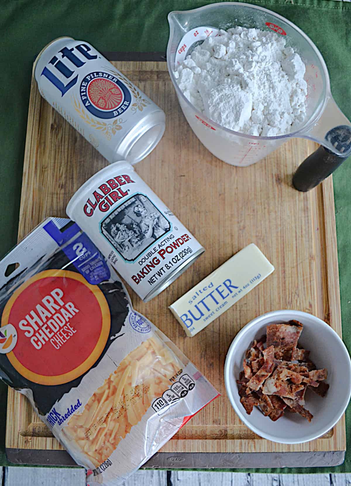 Wooden cutting board with cheese, bacon, a can of beer, butter, and flour on it.