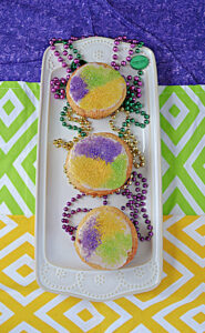 A white platter with 3 cupcakes on it topped with yellow, purple, and green sprinkles and Mardi Gras beads on the platter.