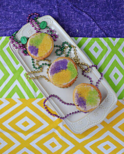 A white platter with 3 cupcakes on it topped with yellow, purple, and green sprinkles and Mardi Gras beads on the platter.