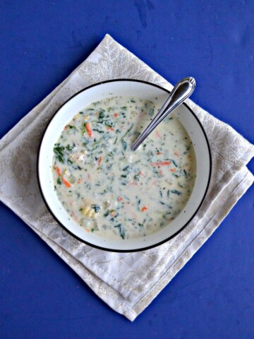 A bowl of creamy white soup dotted with carrots, spinach, and gnocchi with a spoon in it on a white napkin with a blue background.