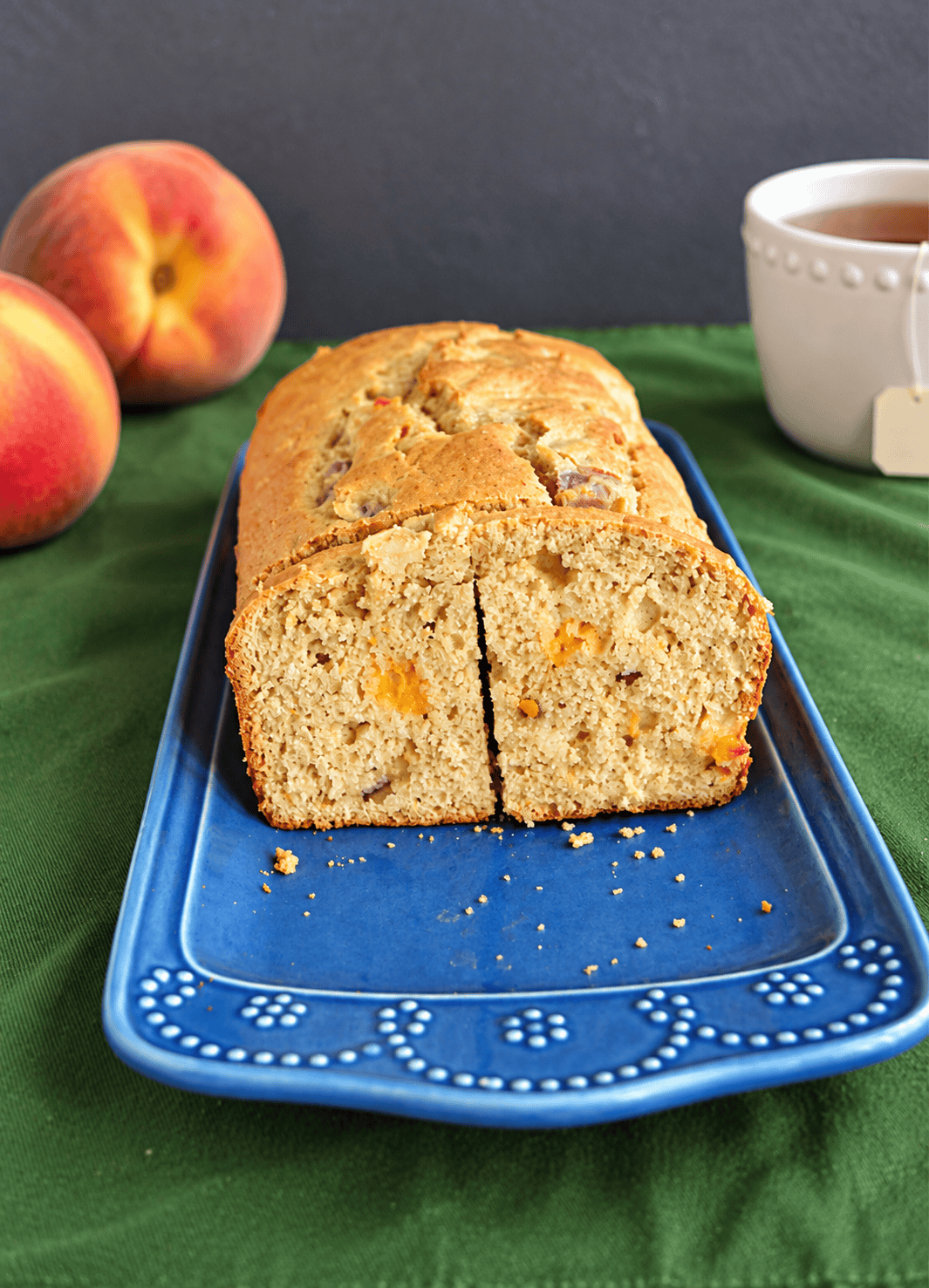 A platter with a loaf of bread on it and 2 peaches and a mug of tea behind the plate.