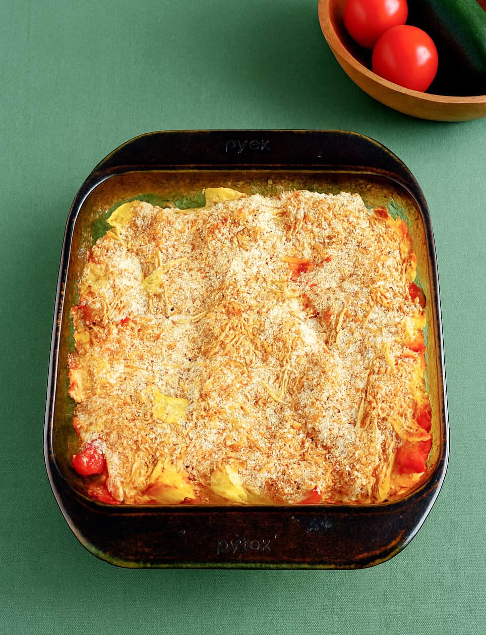 A baking dish full of vegetables and topped off with melted cheese. A bowl of tomatoes and zucchini in the background.