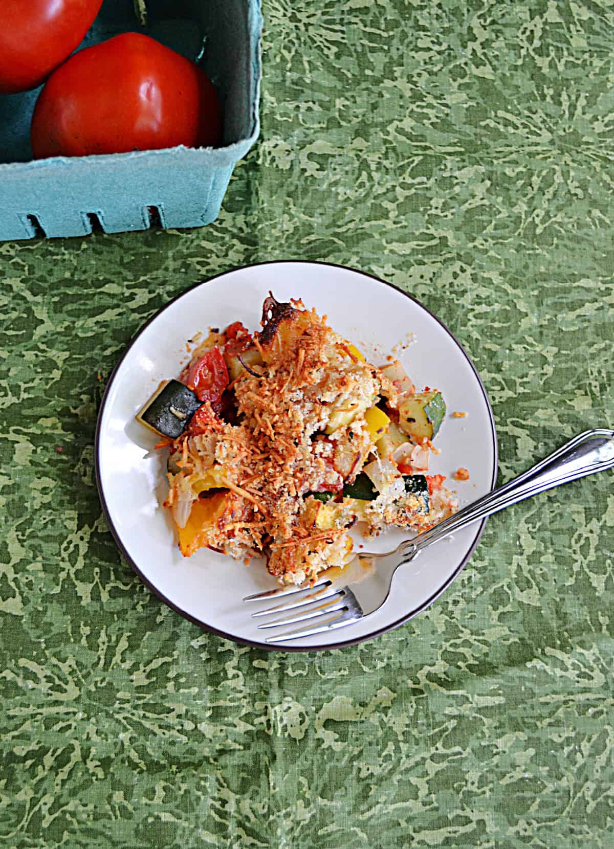 A plate of vegetables topped with cheese with a fork on the plate and a basket of tomatoes in the background.