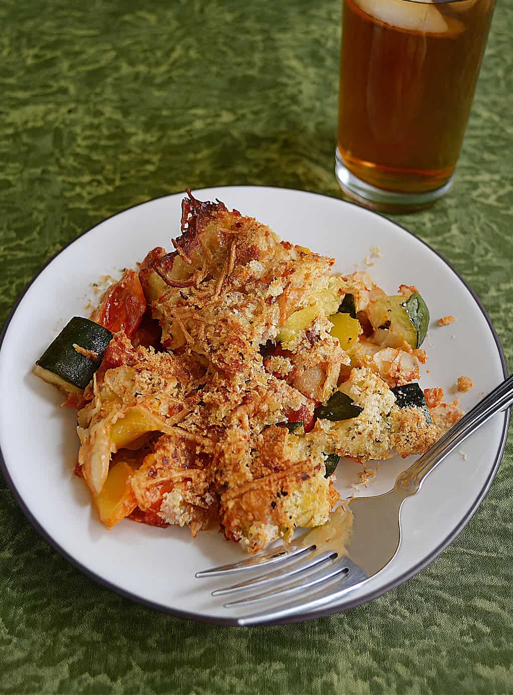 A close up of a plate of vegetables topped with cheese and a fork on the plate with a glass of tea in the background.