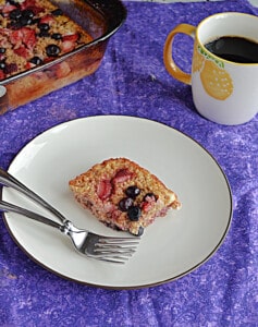 A close up of a plate with a baked oatmeal square on it along with 2 forks, and a cup of coffee behind it.