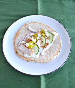Green backdrop with a plate. On the plate is a pita topped with sliced chicken, sliced cucumber, red onions, and feta.