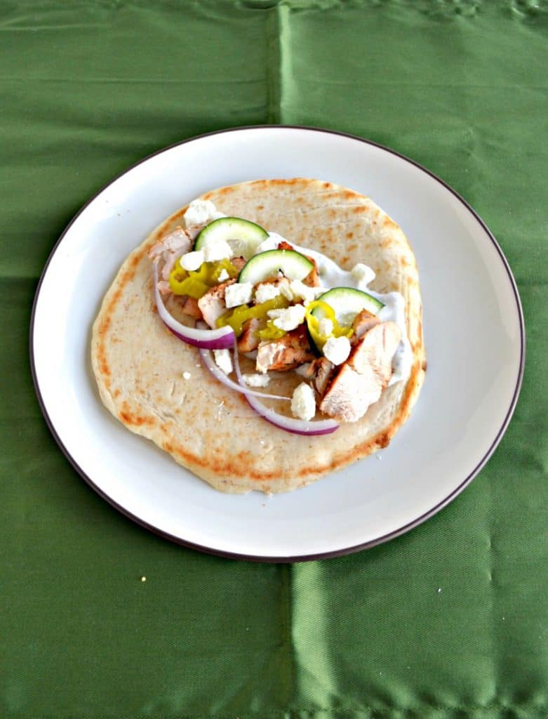 Pita bread on a plate topped with sliced chicken, red onions, feta, and cucumber slices.
