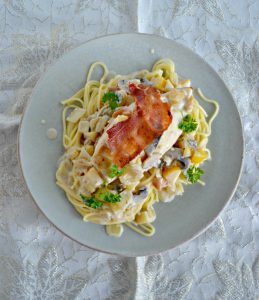 A plate piled high with spagehtti noodles, a creamy sauce, a large chicken breast topped with crispy bacon and fresh parsley on top.