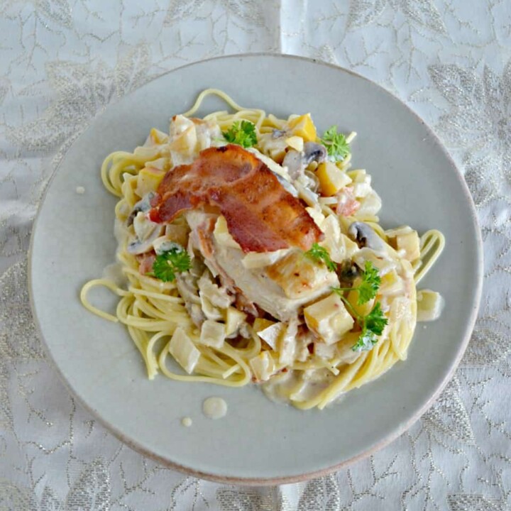 Side view of a plate piled high with spagehtti noodles, a creamy sauce, a large chicken breast topped with crispy bacon and fresh parsley on top.