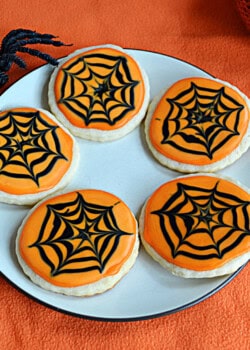 A plate of sugar cookies with orange frosting and black icing spider webs and a spider crawling onto the plate.
