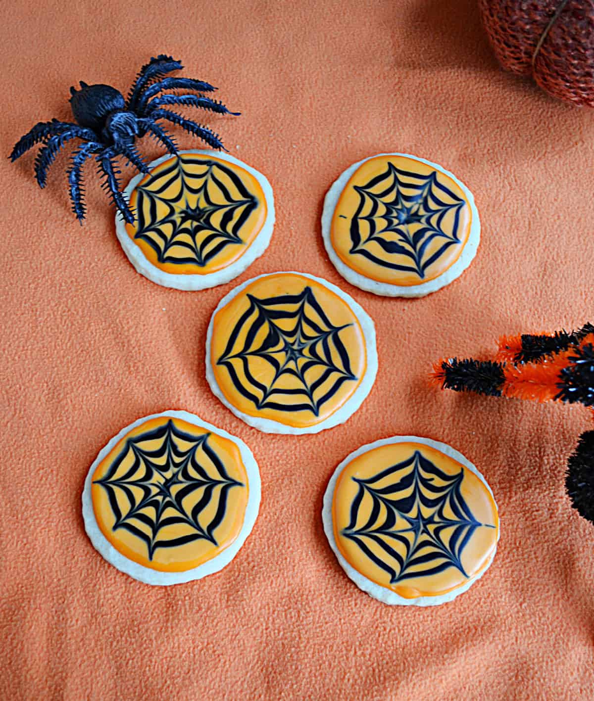A plate of sugar cookies with orange frosting and black icing spider webs and a spider crawling onto the plate.