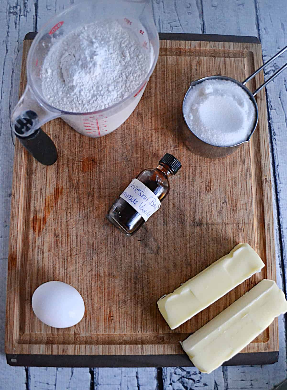 Wooden cutting board with a cup of flour, a cup of sugar, an egg, two sticks of butter, and vanilla extract.