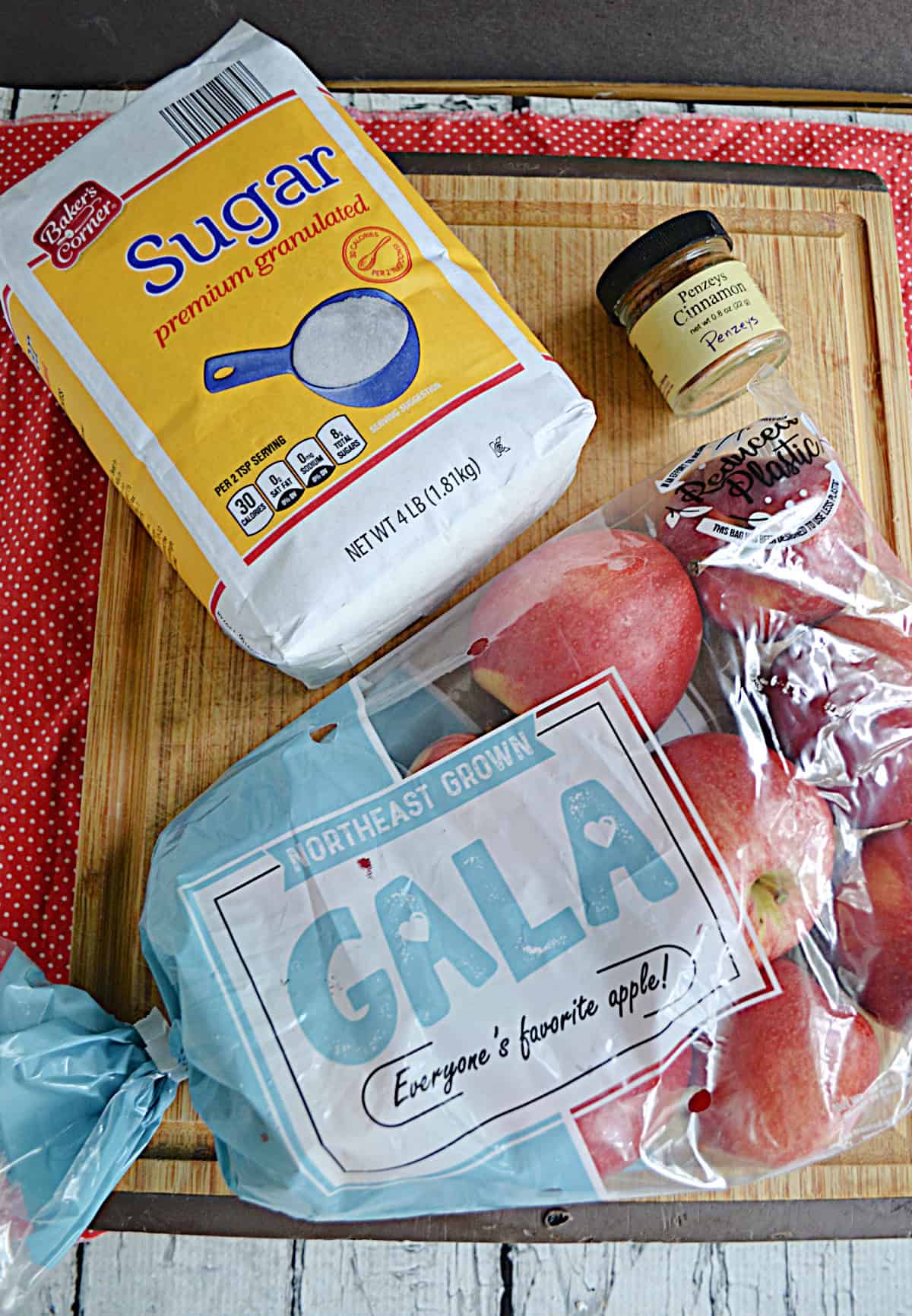 A cutting board with a bag of sugar, a bag of apples, and spices on it.