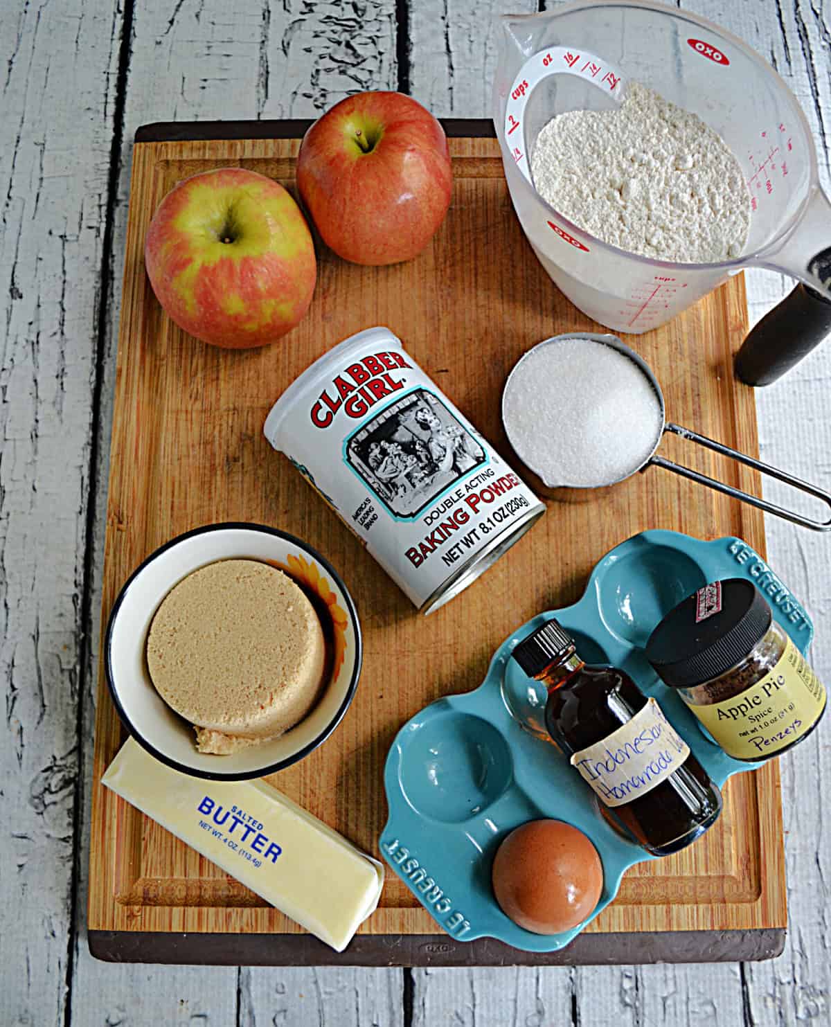 A cutting board with apples, butter, brown sugar, flour, vanilla, eggs, and sugar on it.