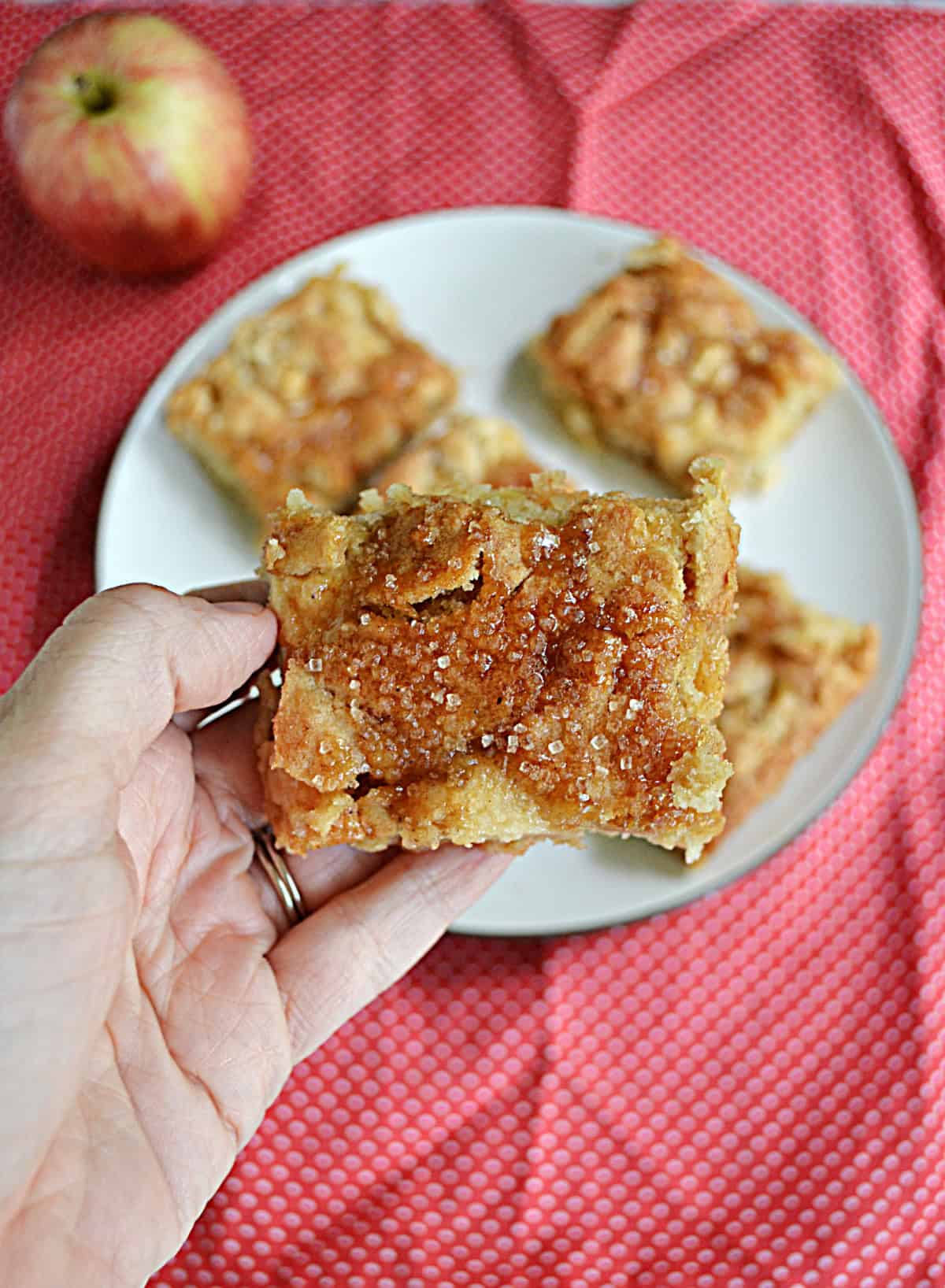 A hand holding an apple square with a plate of apple squares behind.
