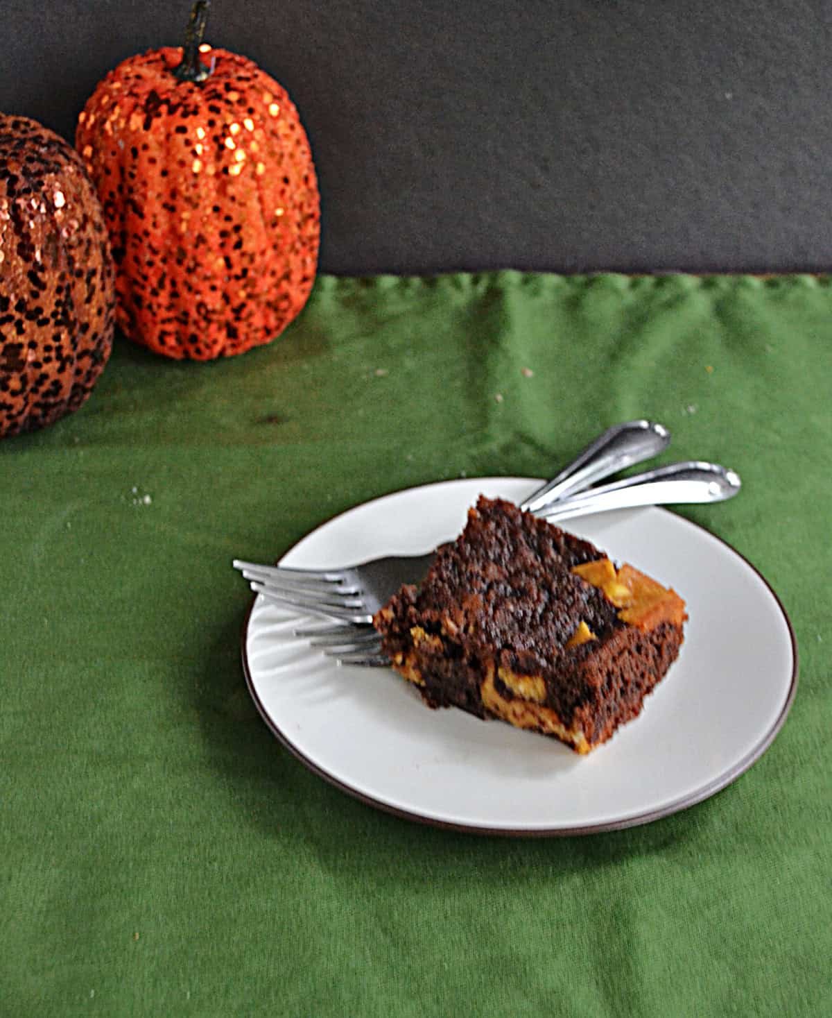 A plate with a pumpkin brownie and 2 forks on it with 2 pumpkins behind it.