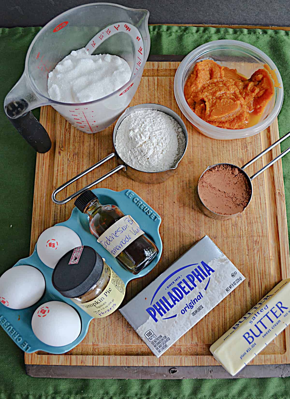 A cutting board with eggs, cream cheese, butter, pumpkin, flour, sugar, and spices.