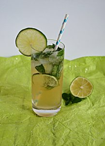 A tall glass with a mojito in it with lime slices and mint leaves floating in it and a straw sstuck in the glass and a half lime beside the glass.
