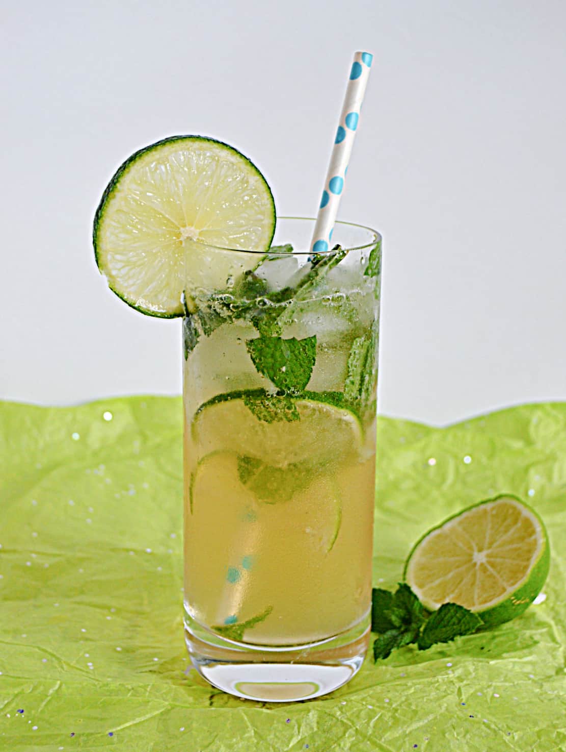 A glass of mojito with a lime slice on the rim and a straw stuck in it and a half lime on the side.